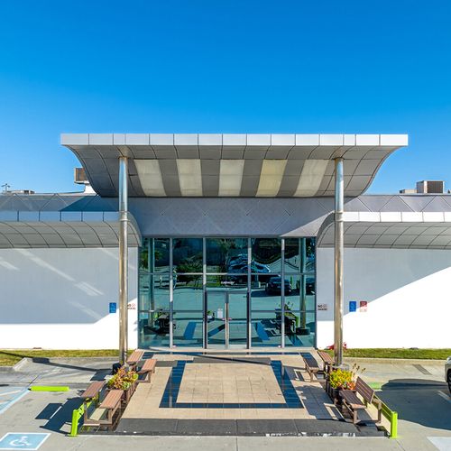 A modern building with large glass doors and a striped canopy sits on a paved area featuring planters and benches The